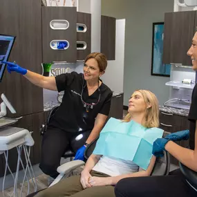 A dentist in black scrubs and blue gloves explains a dental X-ray to a seated patient, pointing at a screen. A dental assistant in black scrubs and gloves observes with a smile. The clinic features modern equipment, dark wood cabinets, and a clean design.