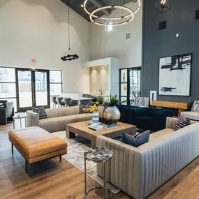 A stylish and modern clubhouse lounge featuring contemporary furniture, high ceilings, and elegant lighting. The space includes a seating area with plush sofas, a coffee table, and accent chairs, as well as a kitchenette and dining area.