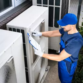 Ein Fachmann in blauer Arbeitskleidung überprüft eine Wärmepumpe in München mit einem Klemmbrett in der Hand. Die Außeneinheiten mehrerer Luft-Wasser-Wärmepumpen stehen an der Hauswand und werden einer Inspektion unterzogen. Die Szene zeigt die Wichtigkeit der Wartung von Wärmepumpen, um deren Effizienz und Langlebigkeit zu gewährleisten.