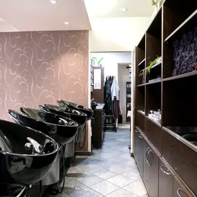 Das Bild zeigt den Waschbereich eines Friseursalons mit einer Reihe von stilvollen, schwarzen Waschbecken, die ergonomisch für eine bequeme Haarwäsche ausgerichtet sind. Die Wand im Hintergrund ist mit einer dekorativen Tapete mit geschwungenem Muster versehen, die dem Raum eine elegante Note verleiht.