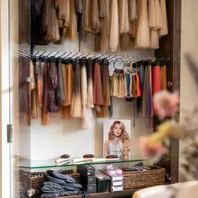 Das Bild zeigt eine sorgfältig organisierte Schauvitrine für Haarverlängerungen in einem Friseursalon. In der oberen Hälfte des Schranks hängen zahlreiche Haarsträhnen in verschiedenen Nuancen – von dunklen Brauntönen bis hin zu hellen Blondtönen – geordnet an Metallhaken. Zusätzlich gibt es eine Auswahl an bunten Extensions in Rot, Blau und weiteren Farben.