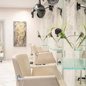 Das Bild zeigt einen eleganten und modern eingerichteten Friseursalon mit einer Reihe von stilvollen Frisierplätzen. Die hellen, beigen Friseurstühle mit verchromten Armlehnen sind vor großen Spiegeln positioniert, die den Raum optisch vergrößern. Die Arbeitsflächen aus Glas verleihen dem Ambiente eine leichte, moderne Note.