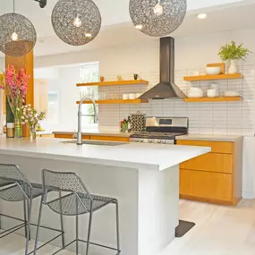 Mid century modern kitchen, tall metal stools along a large kitchen island with sink. large black wire globe light float above. Wood cabinets surround a stove with matching floating shelves over a subway tile back splash.