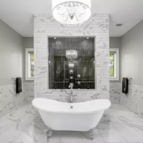 This photo showcases a luxurious bathroom featuring a classic white freestanding clawfoot bathtub as the focal point. The space is adorned with elegant marble tiles on the floor and walls, complemented by a stunning glass-enclosed shower with dark accent tiles. A crystal chandelier hangs above, adding a touch of sophistication and enhancing the spa-like ambiance.