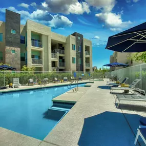 Pool with buildings in the background and patio umbrella up front. Four lounge chairs line the pool.