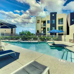 Pool with buildings in the background and patio umbrella up front. Four lounge chairs line the pool.