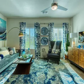 A living area of an apartment with wood floors with a blue patterned rug and a pair of windows with blue patterned curtains