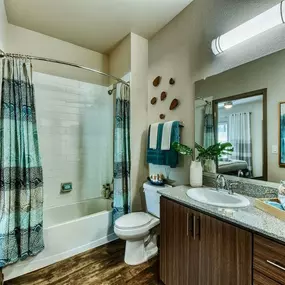 Apartment guest bathroom with white tub and blue shower curtain. Toilet can be seen next to a modern wood vanity and large mirror.