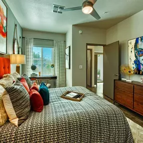 A bedroom with a bed with a grey comforter and a red bed frame. There is a ceiling fan and a window.