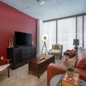 beautifully designed living room in an Atlantic House Midtown apartment, featuring a bold red accent wall, contemporary furnishings, and a cozy yet sophisticated ambiance. The space includes a leather sofa, rustic wooden furniture, and floor-to-ceiling windows that allow for plenty of natural light, creating an inviting and modern living experience in the heart of Atlanta.