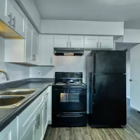 A black refrigerator stands in a kitchen with white cabinets and a stainless steel sink.