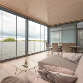 Luxury Waterfront Living Room with Floor-to-Ceiling Glass Views

This elegant waterfront living space showcases expansive floor-to-ceiling glass panels, offering panoramic views of the lake and surrounding nature. The room features light wood flooring and a matching wood-paneled ceiling, creating a warm, coastal-inspired aesthetic. A plush beige sectional sofa with soft accent pillows adds comfort and style, paired with a minimalist coffee table. In the background, a wicker dining set complement