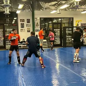 Muay Thai students paired up and practicing striking techniques on blue mats inside a gym, wearing shin guards and gloves, with instructors and spectators watching in the background.