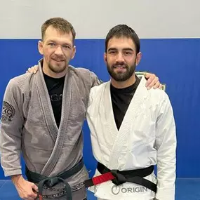 Professor Jordan Blanco and Professor Cory Kuechler, both wearing Brazilian Jiu-Jitsu gis and black belts, posing together on blue mats at a training facility.