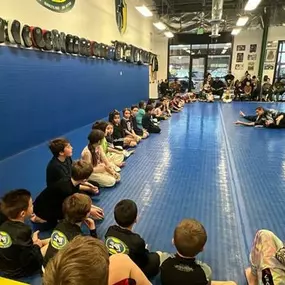Children sitting in a semicircle on blue mats watching a Brazilian Jiu-Jitsu instructor demonstrate a grappling technique inside a gym, with parents and spectators observing from the sidelines.