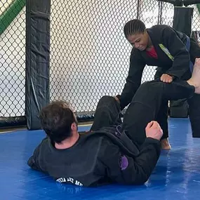 Two Brazilian Jiu-Jitsu practitioners in black gis training on blue mats, with one attempting to pass guard while the other defends inside a fenced training area.