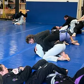 Brazilian Jiu-Jitsu practitioners sparring on blue mats, wearing gis and training in pairs, practicing grappling techniques inside a gym with a fenced training area and shelves with trophies in the background.