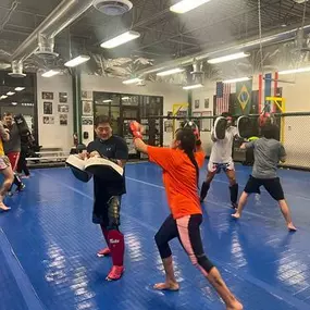 Muay Thai students practicing striking drills with pad holders on blue mats inside Zingano Brighton gym, wearing protective gear, with a fenced training area and international flags in the background.