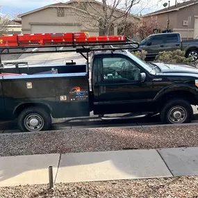 A fully equipped black Perry Mechanical Inc. service truck parked on a residential street, with ladders secured on the roof rack and HVAC tools onboard.
