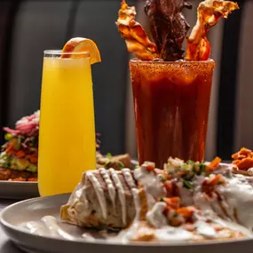 A hearty brunch spread featuring a plate of enchiladas, a bright orange mimosa, and a spicy Bloody Mary crowned with crispy bacon and garnishes, ready to kickstart the day.