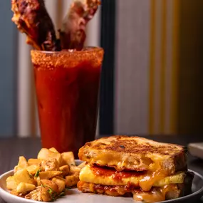 A bold Bloody Mary garnished with crispy bacon and celery sits alongside a plate of stacked grilled golden melted cheese and a side of fresh toppings.