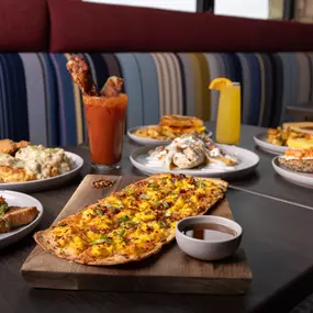 A colorful table setting of loaded breakfast plates, breakfast tacos, and brunch cocktails served in a cozy booth with striped cushions, perfect for a lively meal with friends.