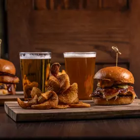 Two hearty burgers with golden onion rings and cold beers sit ready to enjoy, served on a wooden board that showcases this classic comfort food combo.