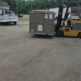 A yellow forklift transports a large HVAC unit in an outdoor parking lot, with service vehicles and a commercial building nearby.