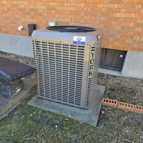 A York outdoor HVAC unit sits on a concrete pad beside a brick house, providing heating and cooling for the building.