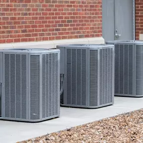 Three large outdoor air conditioning units installed beside a brick building, connected to electrical wiring and refrigerant lines for cooling.