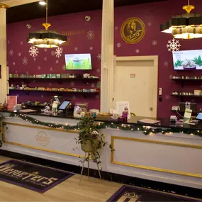 Inside Cannabis Maximus: A welcoming, upscale dispensary in Brattleboro, VT, featuring bold purple-and-gold branding, digital menus, and a fully stocked counter with premium cannabis products.