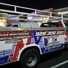 A Nichols Mechanical service truck is parked outside a commercial plaza at night, with storefronts like Crumbl Cookies, uBreakiFix, and Archer’s Bikes visible in the background. The truck features a bold fire-and-ice wrap with prominent branding and service offerings listed on the side, including service maintenance, residential/commercial work, new construction, water treatment service, and jetting & drain cleaning. The phone number and license numbers are clearly displayed, along with a messag