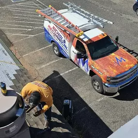 A Nichols Mechanical technician is seen working on a rooftop HVAC or ventilation system, likely part of a commercial electrical or plumbing project in Sedona, AZ. Below, a brightly wrapped Nichols Mechanical service truck is parked, equipped with ladders on its roof. The roof is prepped with Ice & Water Shield underlayment, and shingle work is in progress. The photo captures a clear overhead view of the technician’s precision work, the branded truck, and the surrounding lot.