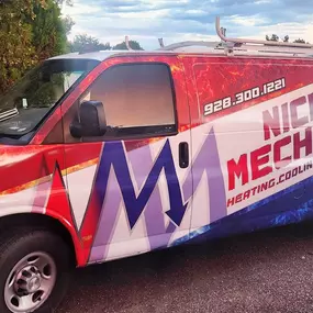 A Nichols Mechanical service van is parked on a gravel lot beside a large tree, showcasing the company’s bold, full-vehicle wrap. The design features striking red and blue graphics with fire and ice elements, representing their HVAC expertise. Prominently displayed on the van are the company name, phone number, and service areas: heating, cooling, plumbing, and electrical. A ladder rack is mounted on the roof, and the sky is softly lit with evening clouds in the background.