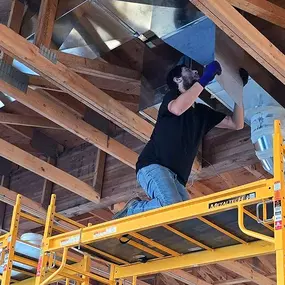 A technician from Nichols Mechanical is working on ductwork installation in a commercial building under construction. He is kneeling on a yellow scaffold while connecting or inspecting a large metal HVAC duct attached to the wooden trusses above. The open framing and exposed structure highlight the early stage of the build, with visible support beams and ventilation lines in progress.