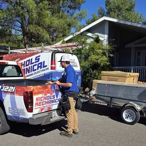 Two technicians from Nichols Mechanical are on-site in front of a residential home, preparing for an HVAC installation. One technician is working at the back of a branded Nichols Mechanical pickup truck, while the other is handling equipment on a trailer hitched to a Nichols Mechanical service van. The vehicles are parked in a shaded neighborhood with pine trees and a covered porch visible in the background. The company’s branding and contact information are clearly displayed on the vehicles, em