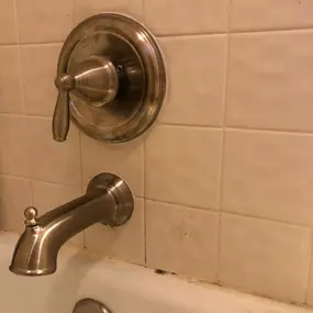 This image displays a bathtub faucet and shower control handle with a brushed nickel finish against a white tiled wall. Visible signs of aging include discolored grout and minor dirt buildup near the tub’s edge.