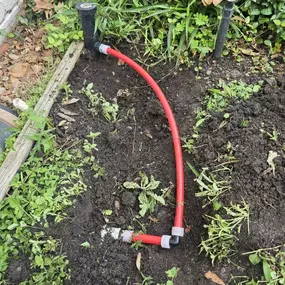 This image showcases a sprinkler system repair featuring a red PEX pipe with elbow fittings connecting to a sprinkler head. The work is done in a garden bed with exposed soil, plants, and irrigation components.