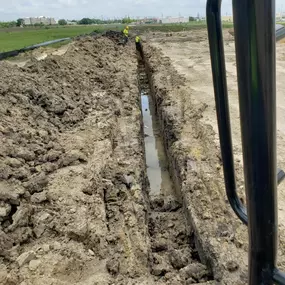 The image shows a large construction site with a long, narrow trench filled with water. Two workers wearing safety gear stand in the trench, while an excavator's controls are visible in the foreground.