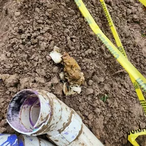 The image captures a muddy construction area with broken PVC pipes, a rotting organic object on the dirt, and yellow caution tape stretched nearby, indicating a possible hazard or repair work.