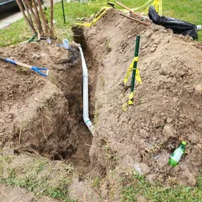 The image shows an excavation site in a grassy yard with a deep trench exposing a newly installed PVC pipe. Yellow caution tape, digging tools, and scattered debris indicate an ongoing underground utility repair.