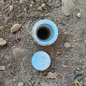 The image shows an open blue PVC pipe embedded in dry, rocky soil, with its cap placed beside it. A wrench is partially visible in the lower right, suggesting maintenance or inspection work.