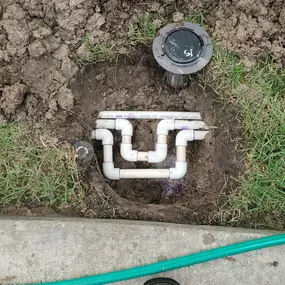 The image shows an exposed underground PVC irrigation system with interconnected pipes and fittings in muddy soil. A black valve box is nearby, with a green hose and a worker's boot next to a concrete sidewalk.