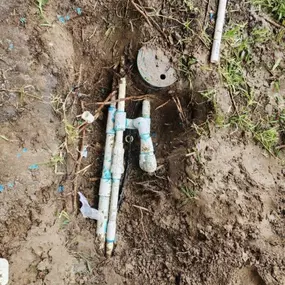 The image shows an exposed irrigation system with multiple PVC pipes, some wrapped in blue tape and appearing damaged or broken. The surrounding soil is muddy, with grass patches and scattered debris nearby.