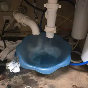 The image captures a plumbing issue under a sink where a disconnected PVC pipe is leaking water into a blue plastic bowl. Surrounding elements include rags, wires, and various plumbing components.