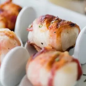 Golden, crispy bacon-wrapped scallops garnished with herbs, presented in ceramic serving spoons on a white tray.