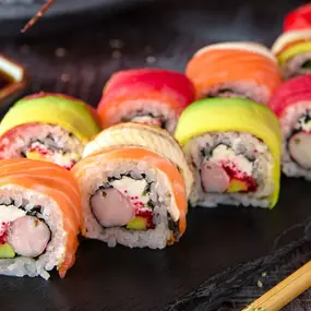 Colorful rainbow sushi rolls filled with crab, avocado, and cream cheese, topped with fresh slices of salmon, tuna, and avocado, arranged on a black slate with chopsticks and soy sauce.