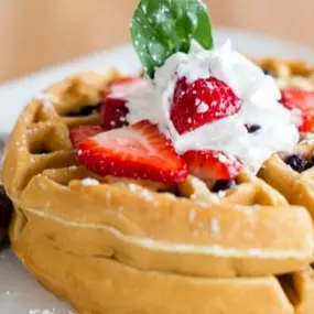 Fluffy Belgian waffles topped with whipped cream, fresh strawberries, and a mint leaf, served with sausage links on a white plate from our Breakfast Menu