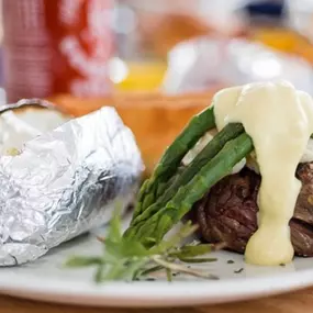 Grilled filet mignon topped with steamed asparagus and creamy hollandaise sauce, served with a foil-wrapped baked potato on a white plate at Miller's Seafood & Steakhouse in Kill Devil Hills, NC.