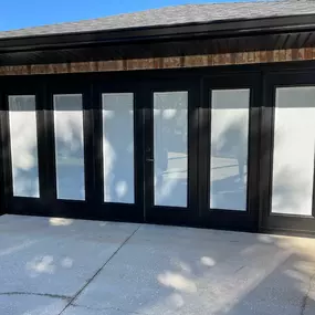 This image shows a newly installed or painted set of six black garage doors with frosted or blinded windows, set into a brick wall. A concrete driveway extends from the doors under a clear sky.
